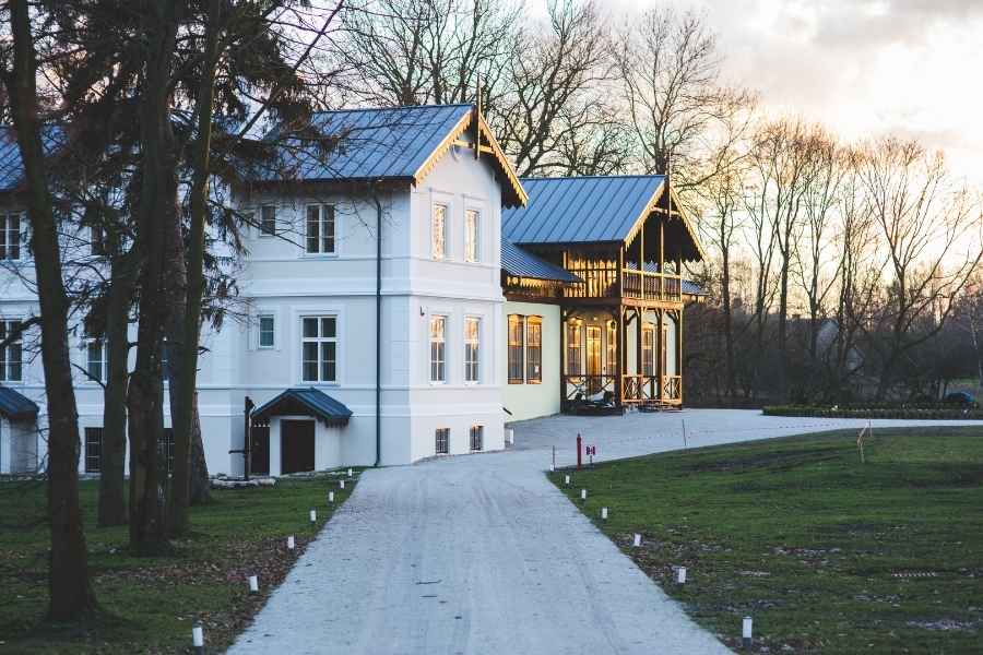 Une grande maison blanche avec toiture en métal et véranda en bois illuminée au crépuscule, représentant un bien immobilier de caractère en milieu naturel.