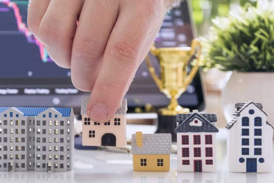 Main choisissant une maquette de maison parmi plusieurs avec un trophée et des graphiques boursiers pour symboliser le premier investissement immobilier
