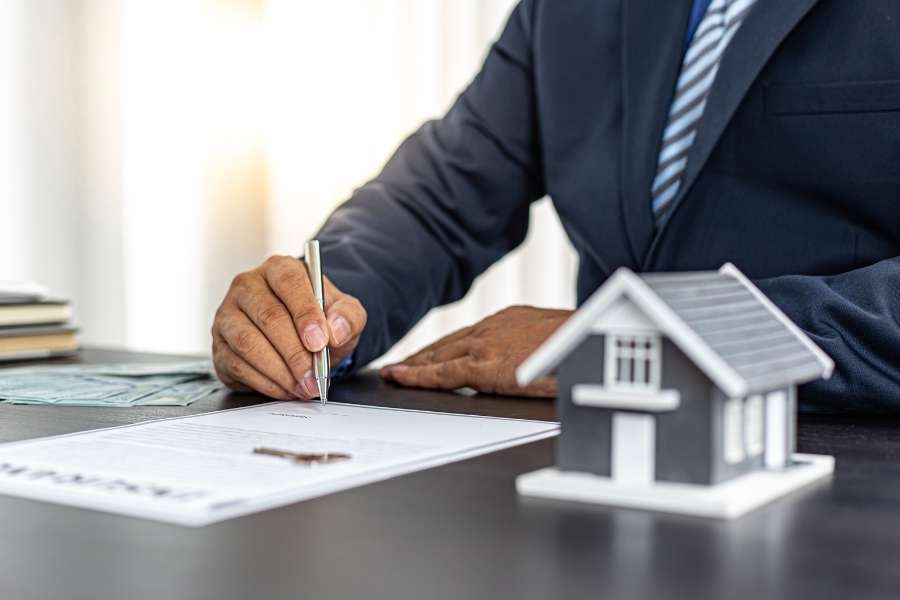 Un homme en costume signe un contrat à côté d'une maquette de maison pour l'acquisition de biens immobiliers.